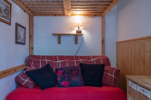 Une pièce chaleureuse avec un plafond en bois, un canapé rouge agrémenté de coussins à motifs, une lampe fixée au mur et des tableaux encadrés.