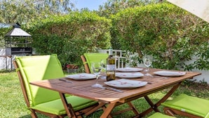Un área de comedor al aire libre con una mesa de madera dispuesta para cuatro personas, rodeada de sillas verdes, una botella de vino y una copa.