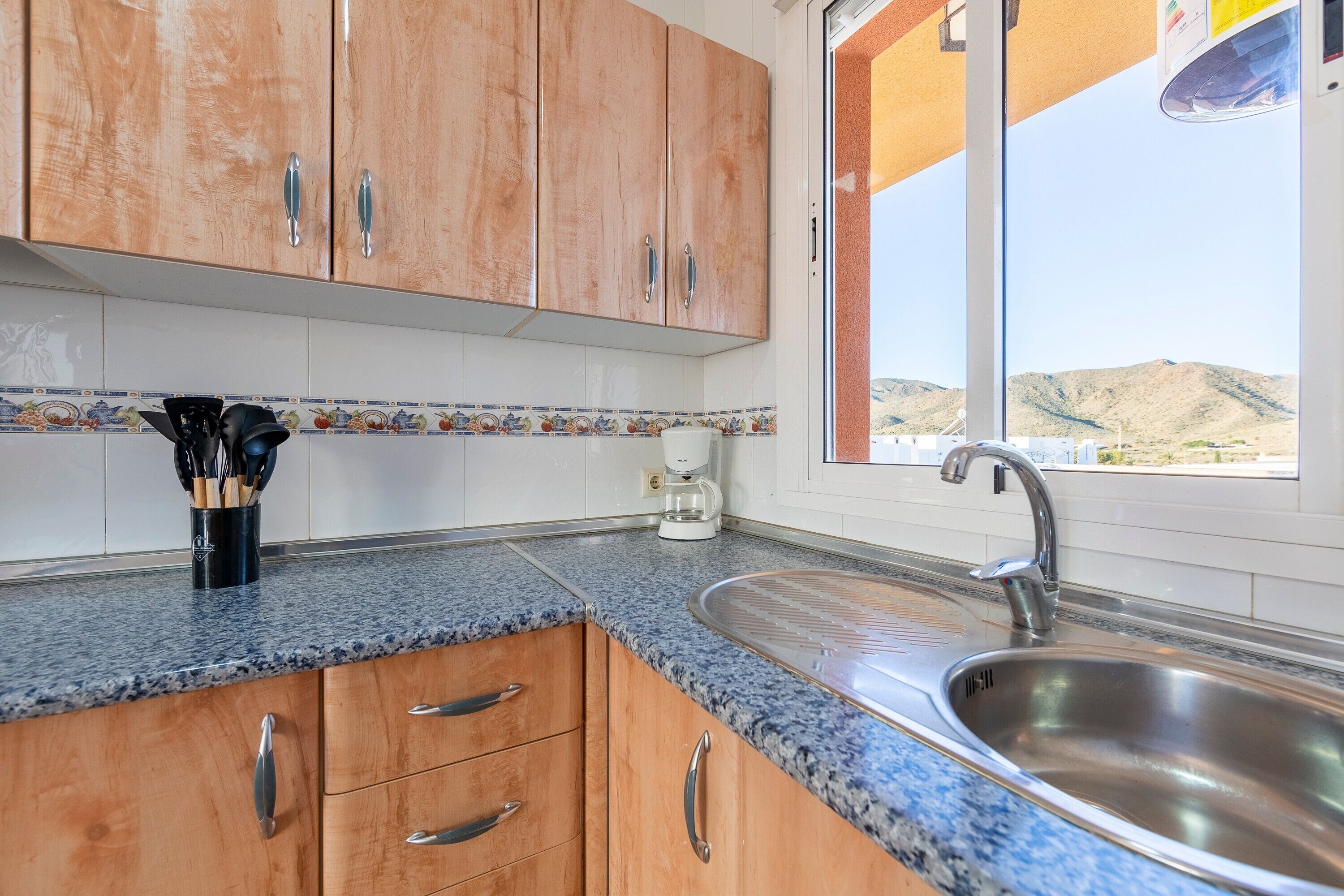 A kitchen with wooden cabinets, a granite countertop, a stainless steel sink, and a window with a view of hills.