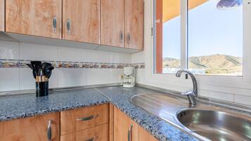 A kitchen with wooden cabinets, a granite countertop, a stainless steel sink, and a window with a view of hills.