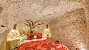 Une pièce avec un mur en pierre, un lit recouvert d’une couverture à motifs, deux oreillers, un vase avec des branches et des lampes fixées au mur.