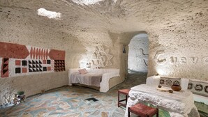 Une pièce qui ressemble à une grotte, avec un sol en pierre, un lit, une table avec des tabourets et des décorations murales.
