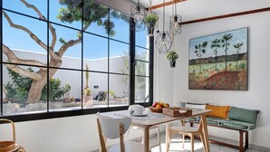 Un área de comedor con una mesa, sillas, un banco y un ventanal con vistas a árboles y un cuadro de paisaje.