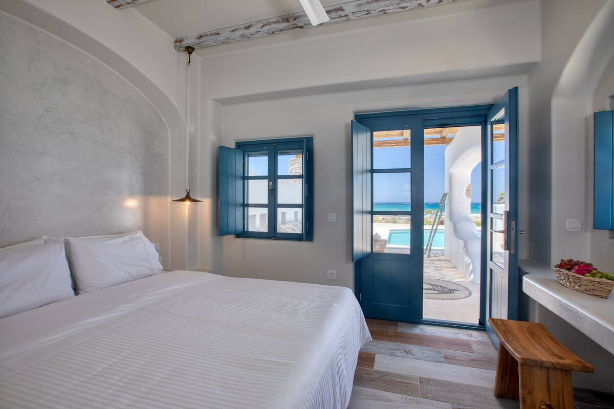 A bedroom with a bed, a wooden bench, a door leading to a balcony, and a view of the sea.