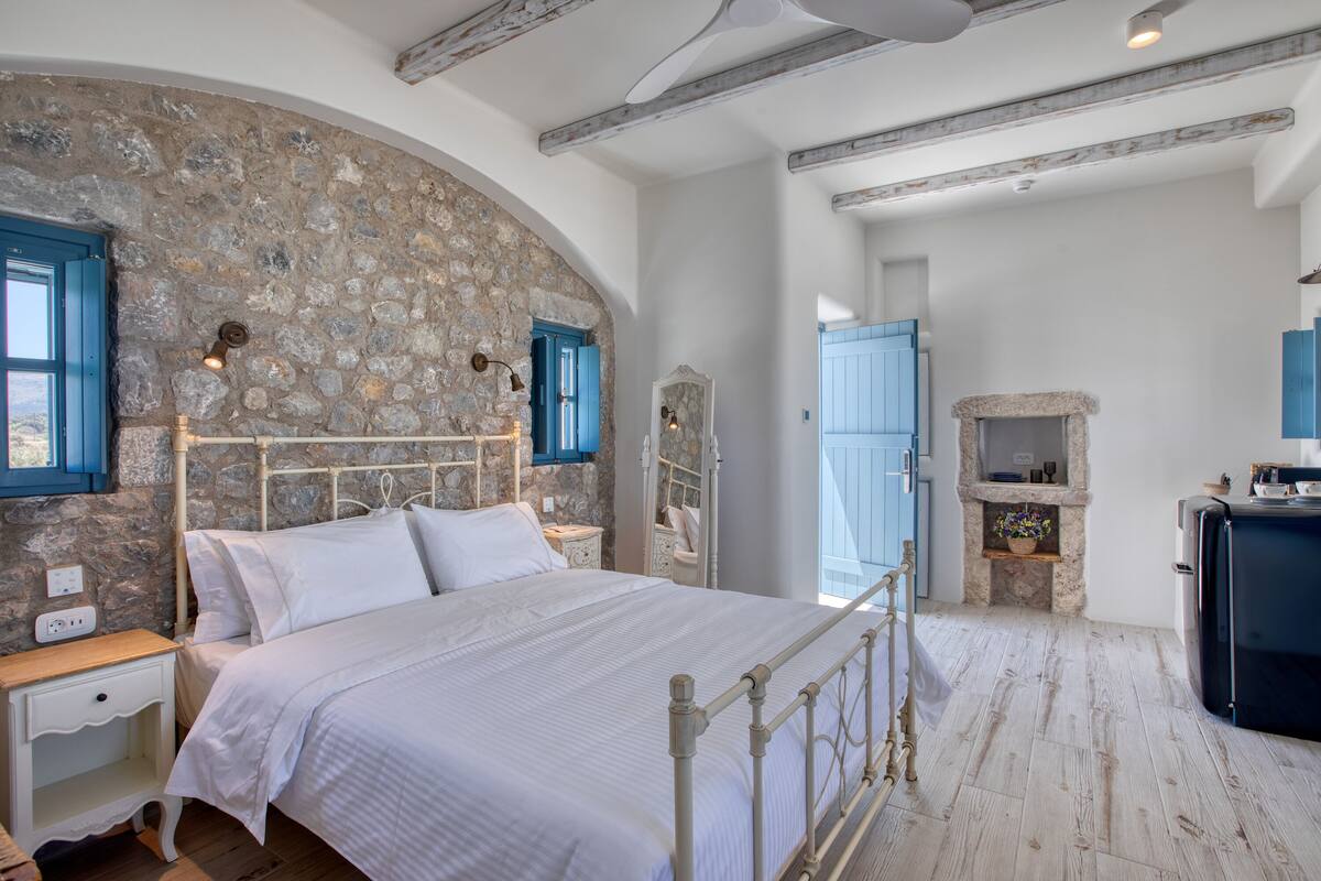 A bedroom with a stone wall, a bed with white linens, a wooden nightstand, a dresser, and a small stone fireplace.