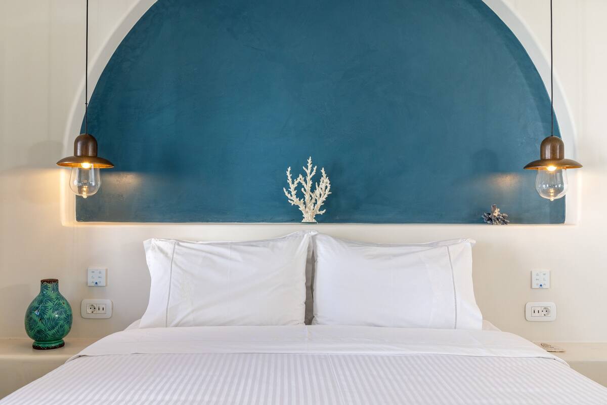 A neatly made bed with white linens, a white vase on the nightstand, and a blue wall arch above.