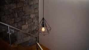 Eine sechseckige Pendelleuchte mit sichtbarer Glühbirne, die von der Decke in einem Raum mit Steinwand und Holztreppe hängt.