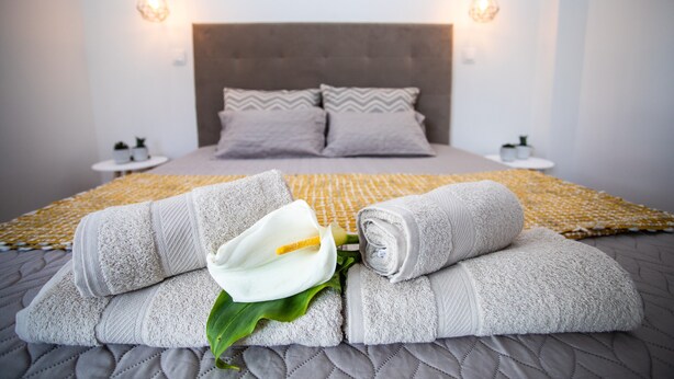 Ein ordentlich bezogenes Bett mit aufgerollten Handtüchern, einer weißen Blume, zwei Nachttischen und einem Kopfteil.