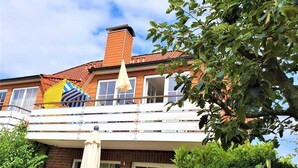 Ein zweistöckiges Haus mit Balkon, Schornstein und einem Baum im Vordergrund.