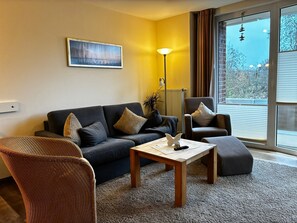 Een moderne woonkamer met een bank, fauteuil en salontafel. Er hangt een schilderij aan de muur, er staat een vloerlamp en er is een raam met zonwering.