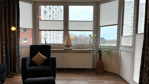 A room with a black armchair, a wooden floor, a radiator, a window with blinds, and a view of buildings and trees. - TRA-152 - Residence Trafalgar (Cuxhaven)
