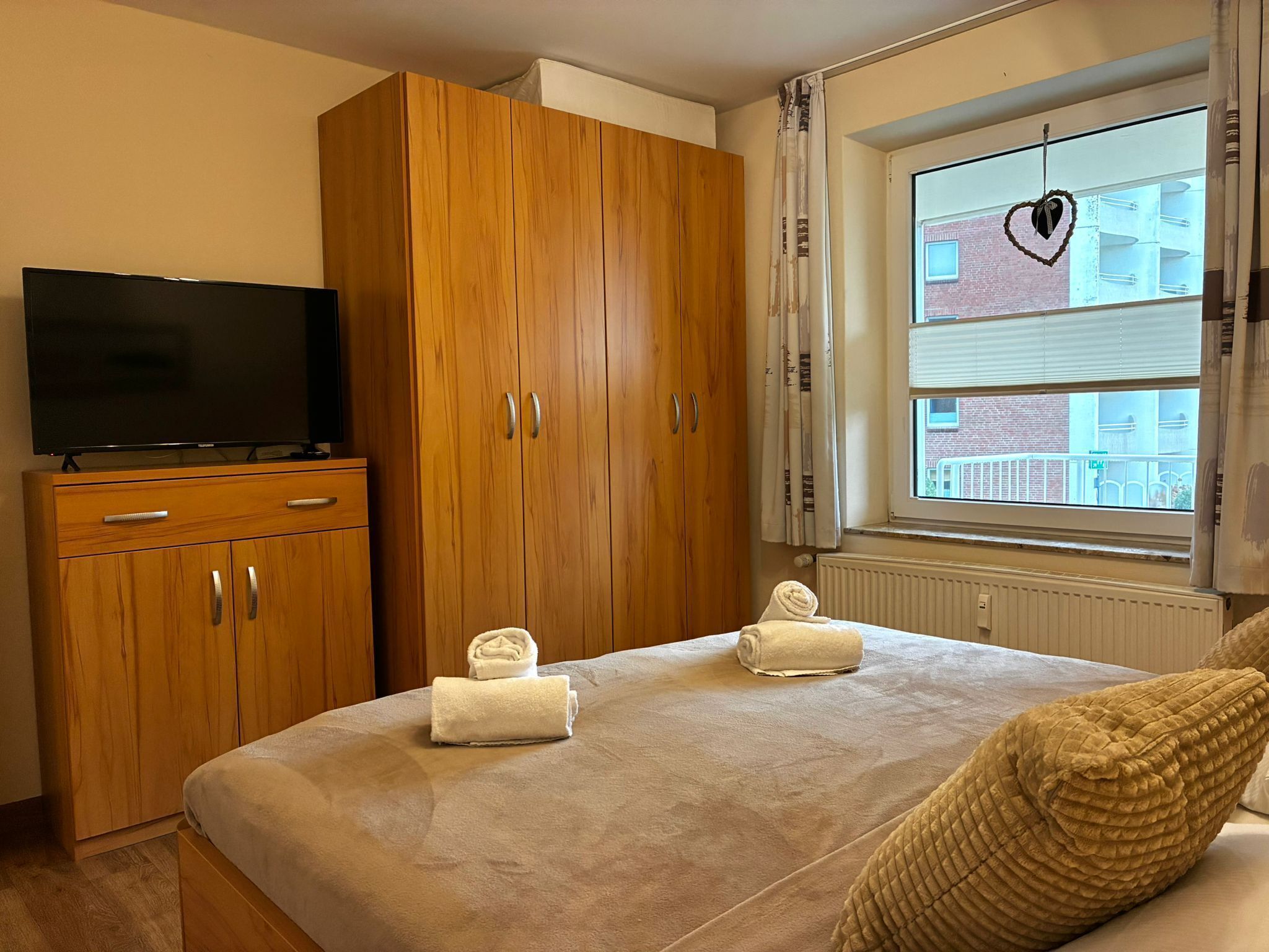 Une chambre à coucher avec un lit, un téléviseur posé sur un meuble en bois et une fenêtre donnant sur des immeubles.