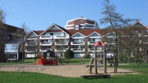 A playground with swings and a climbing structure in front of a building with multiple balconies and a red roof. - TRA-230 - Residence Trafalgar (Cuxhaven)
