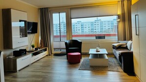 A modern living room with a sofa, a coffee table, a TV, and a large window with curtains. - TRA-431 - Residence Trafalgar (Cuxhaven)