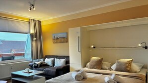 A modern hotel room with a bed, a sofa, a coffee table, and a view of the outside. - TRA-431 - Residence Trafalgar (Cuxhaven)