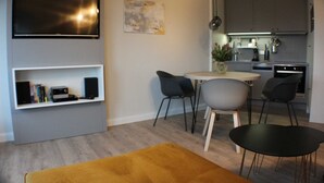 A modern living room with a flat-screen TV, a bookshelf, a dining area with a table and chairs, and a kitchen in the background. - TRA-525 - Residence Trafalgar (Cuxhaven)