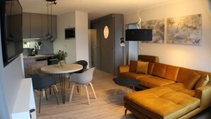A modern living room with a sofa, a dining table with chairs, and a kitchen area in the background. - TRA-525 - Residence Trafalgar (Cuxhaven)