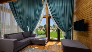 A modern living room with a grey sectional sofa, a grey ottoman, and a large window with teal curtains. - Villa d'Mare (Demre)