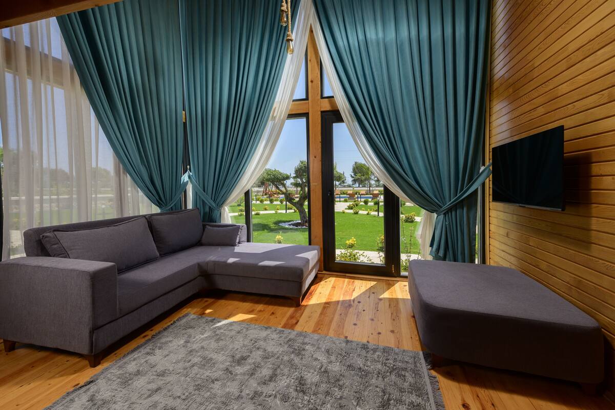 A modern living room with a grey sectional sofa, a grey ottoman, and a large window with teal curtains.