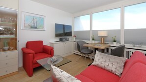 Un salon moderne avec un canapé rouge, un fauteuil rouge, une table basse en verre, un téléviseur à écran plat et un coin repas comprenant une petite table et des chaises.