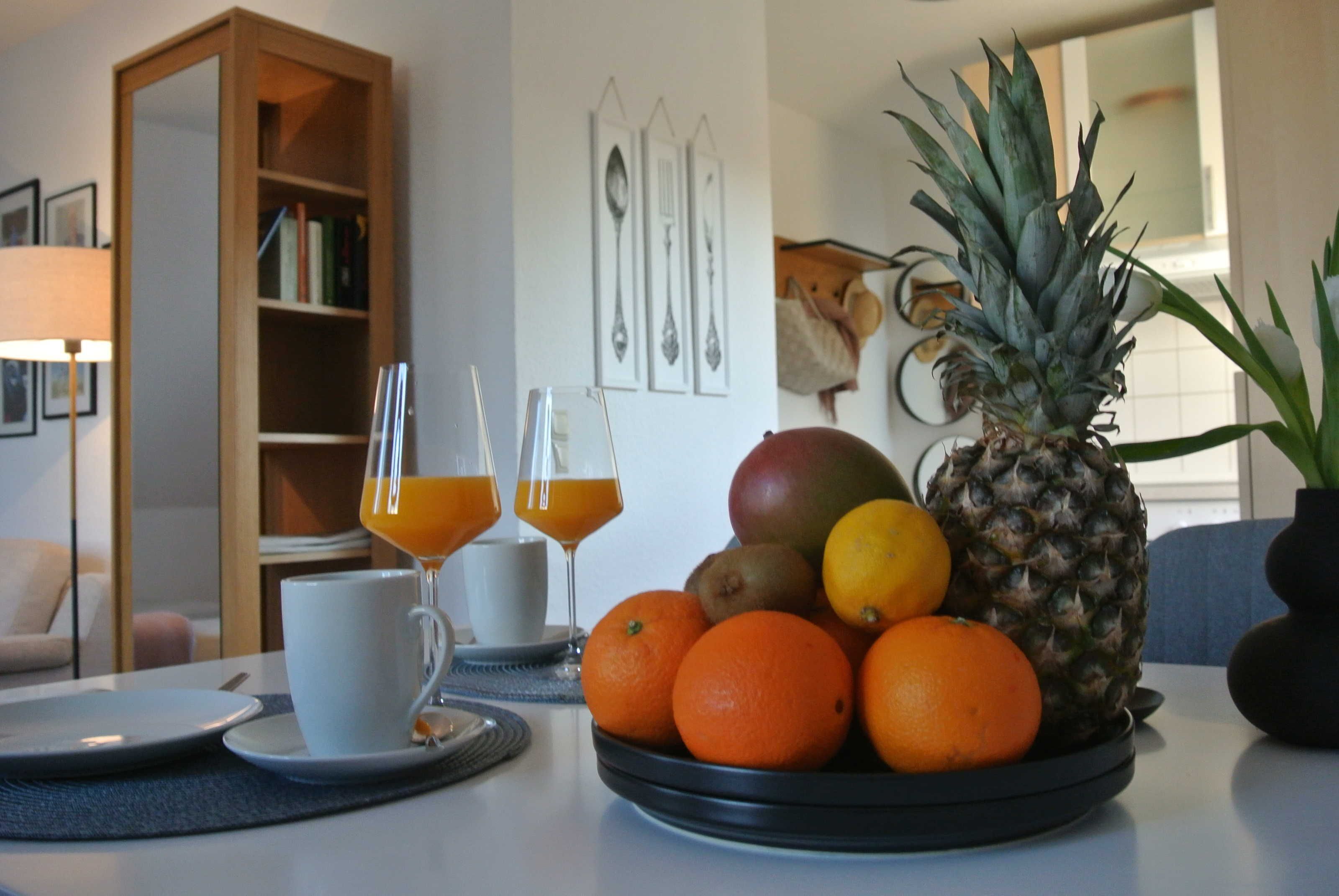 Una tavola per la colazione con una ciotola di frutta contenente arance, un'ananas e un mango. Si vedono due bicchieri di succo d'arancia e una tazza bianca sul tavolo.