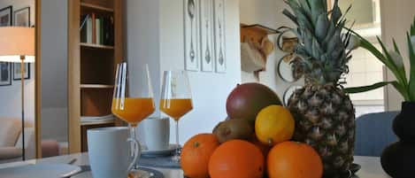 A breakfast table with a fruit bowl containing oranges, a pineapple, and a mango. There are two glasses of orange juice and a white cup on the table.