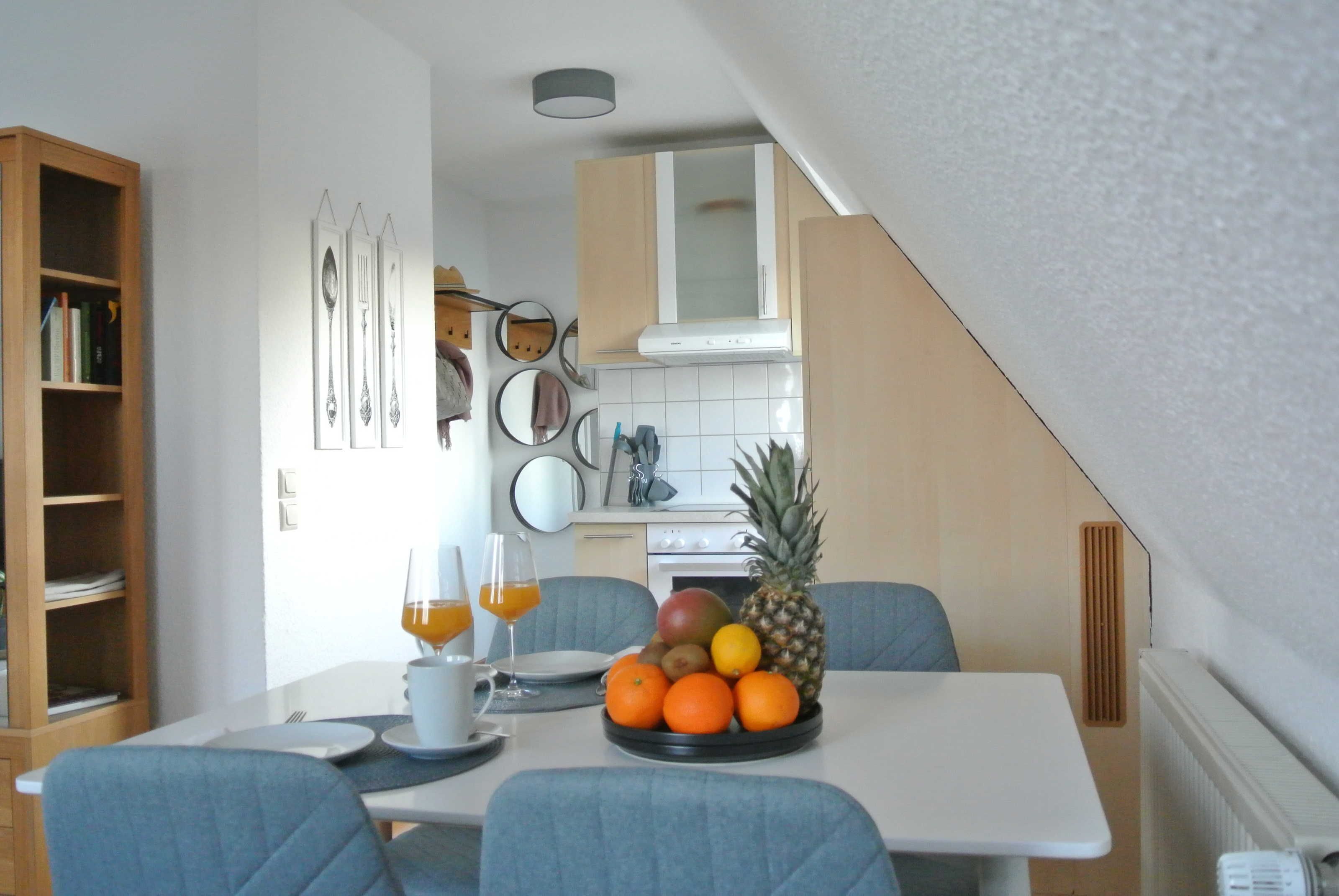 A compact kitchen with a dining table set for two, featuring a fruit bowl with oranges and a pineapple.