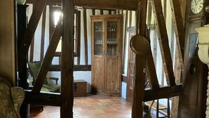 Ein rustikales Interieur mit freiliegenden Holzbalken, einem Fischgrätparkett und antiken Möbeln, darunter ein mit Grünstoff gepolsterter Sessel und ein Holzschrank mit Glastüren.