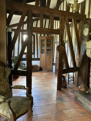 Ein rustikales Interieur mit freiliegenden Holzbalken, einem Fischgrätparkett und antiken Möbeln, darunter ein mit Grünstoff gepolsterter Sessel und ein Holzschrank mit Glastüren.