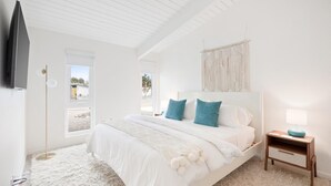 A bedroom with a large bed, a TV mounted on the wall, a bedside table with a lamp, and a macrame wall hanging.