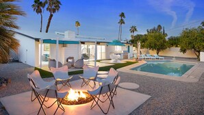 A modern outdoor pool area with a fire pit and lounge chairs, surrounded by palm trees and a clear sky.