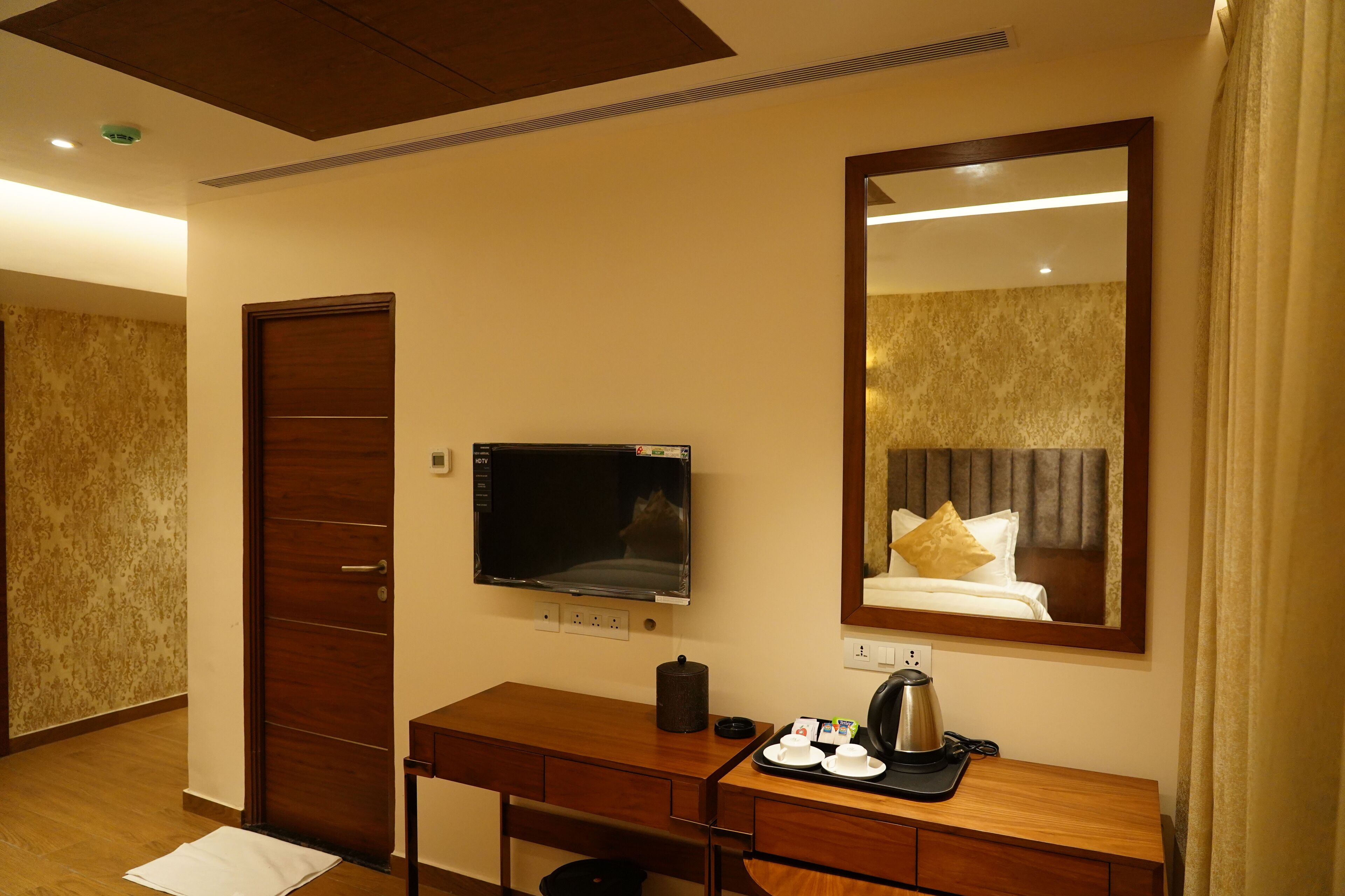 A hotel room with a wooden desk, a flat-screen TV, a bed with a headboard, and a large mirror.