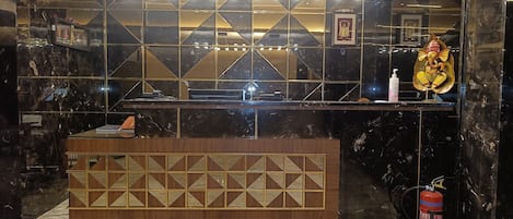 Bancada de recepção com piso de mármore, um extintor de incêndio e uma parede decorativa com padrões geométricos.