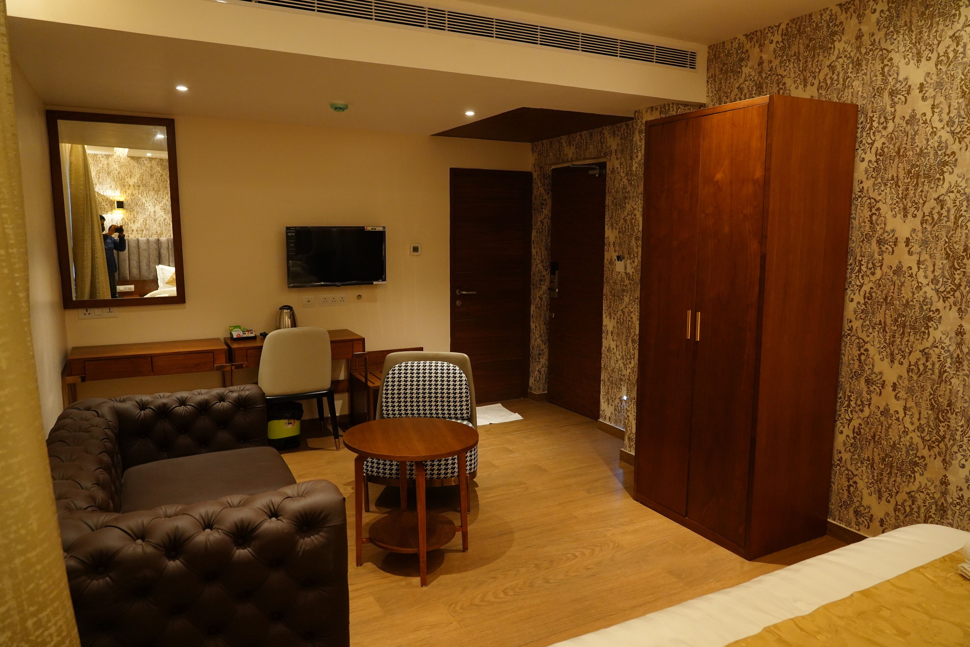 A hotel room with a sofa, a small table, a chair, a wardrobe, and a TV.