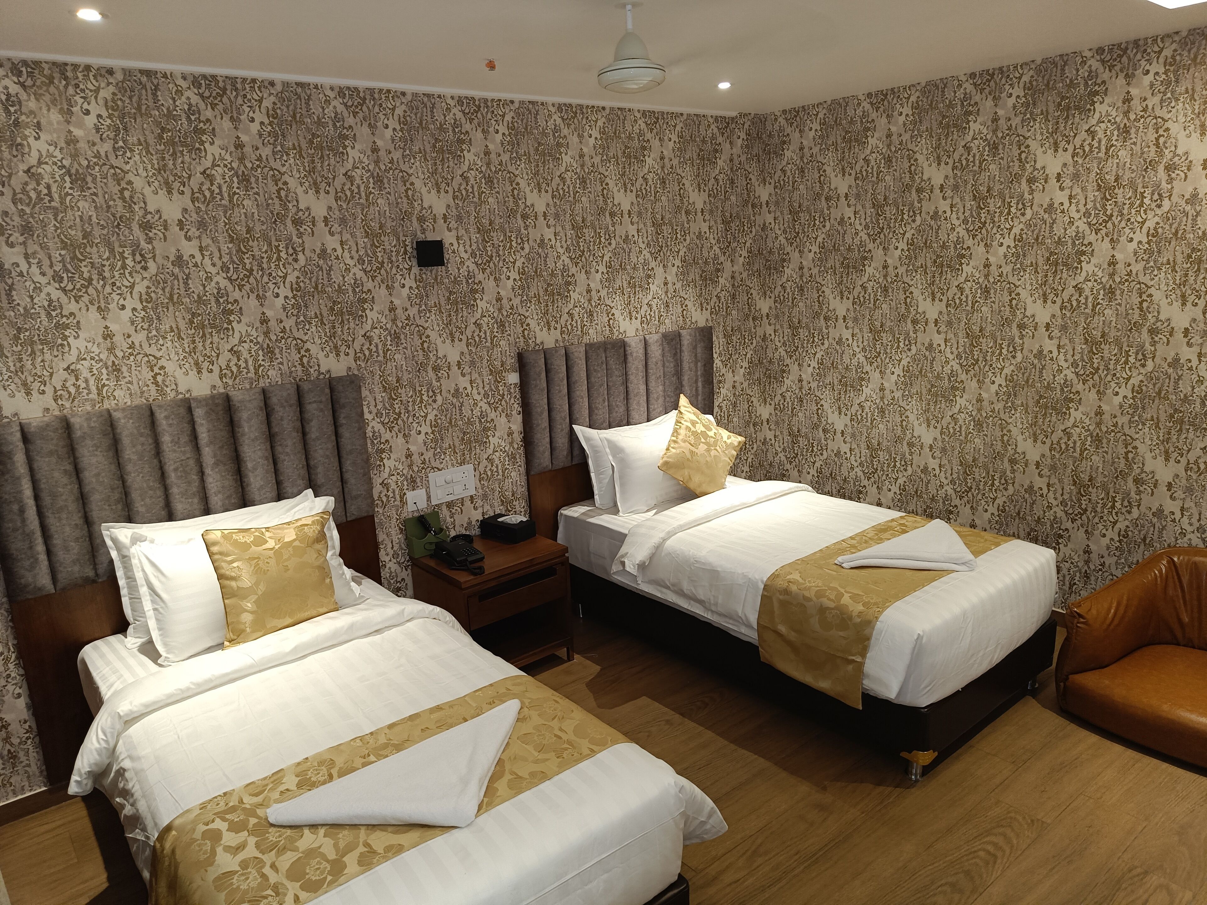 Une chambre d’hôtel avec deux lits, chacun doté d’une tête de lit, d’une table de chevet et d’un téléphone. La chambre est décorée avec un papier peint à motifs et comprend un fauteuil en cuir marron.