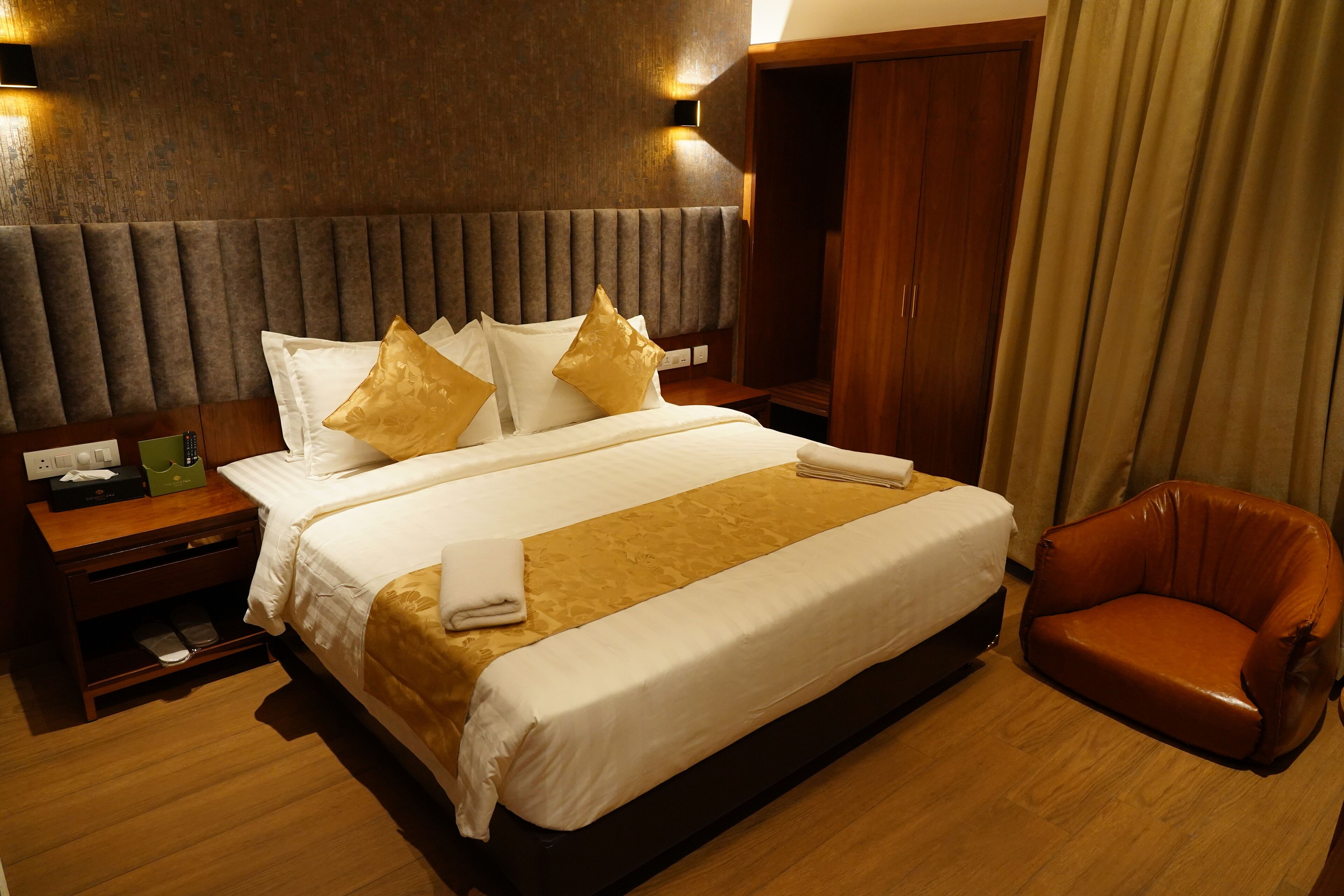 Una habitación de hotel con una cama grande, un cabecero de madera, un sillón de cuero marrón y una mesita de noche de madera con una lámpara.