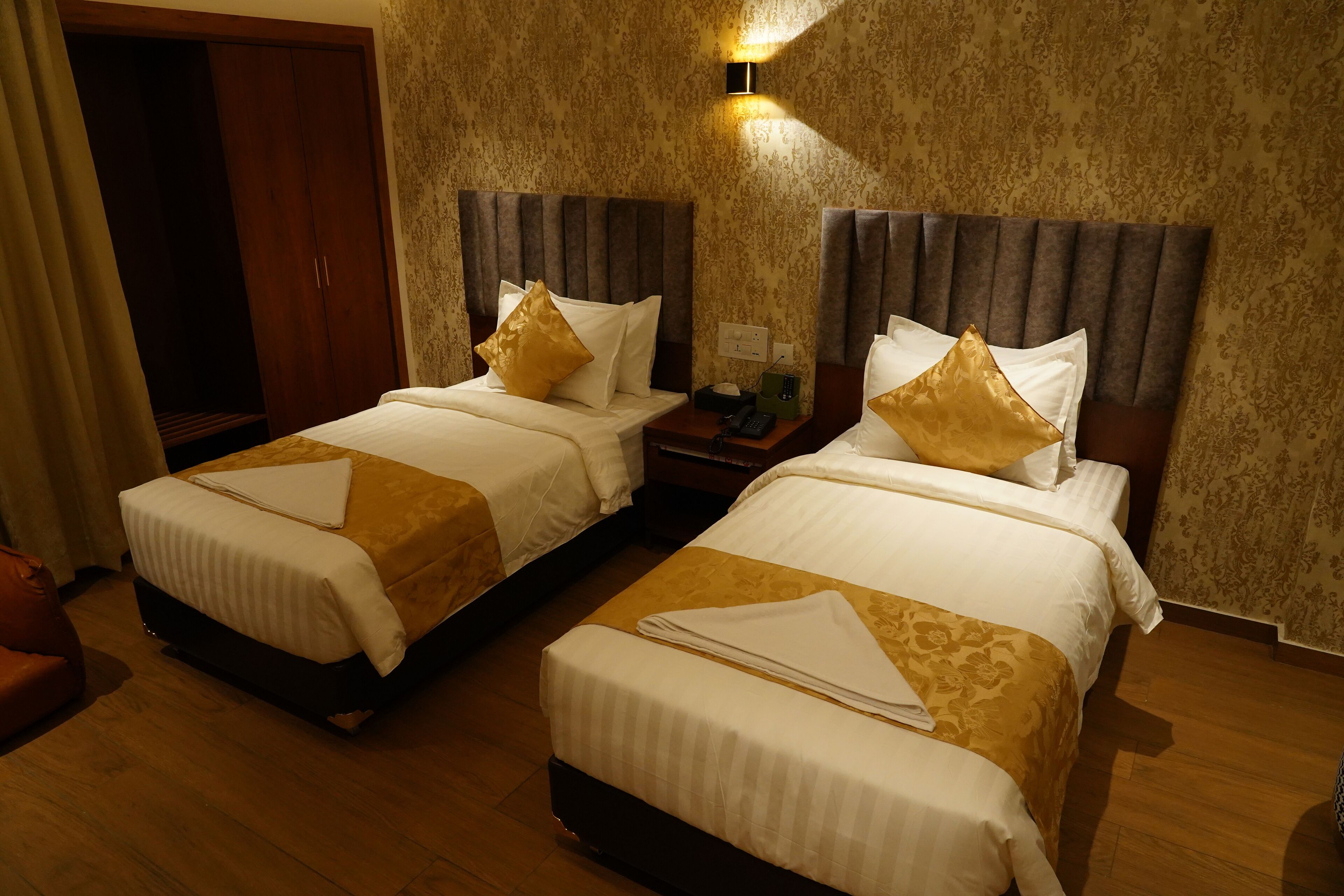 A hotel room with two beds, wooden flooring, a wardrobe, and a nightstand with a phone.