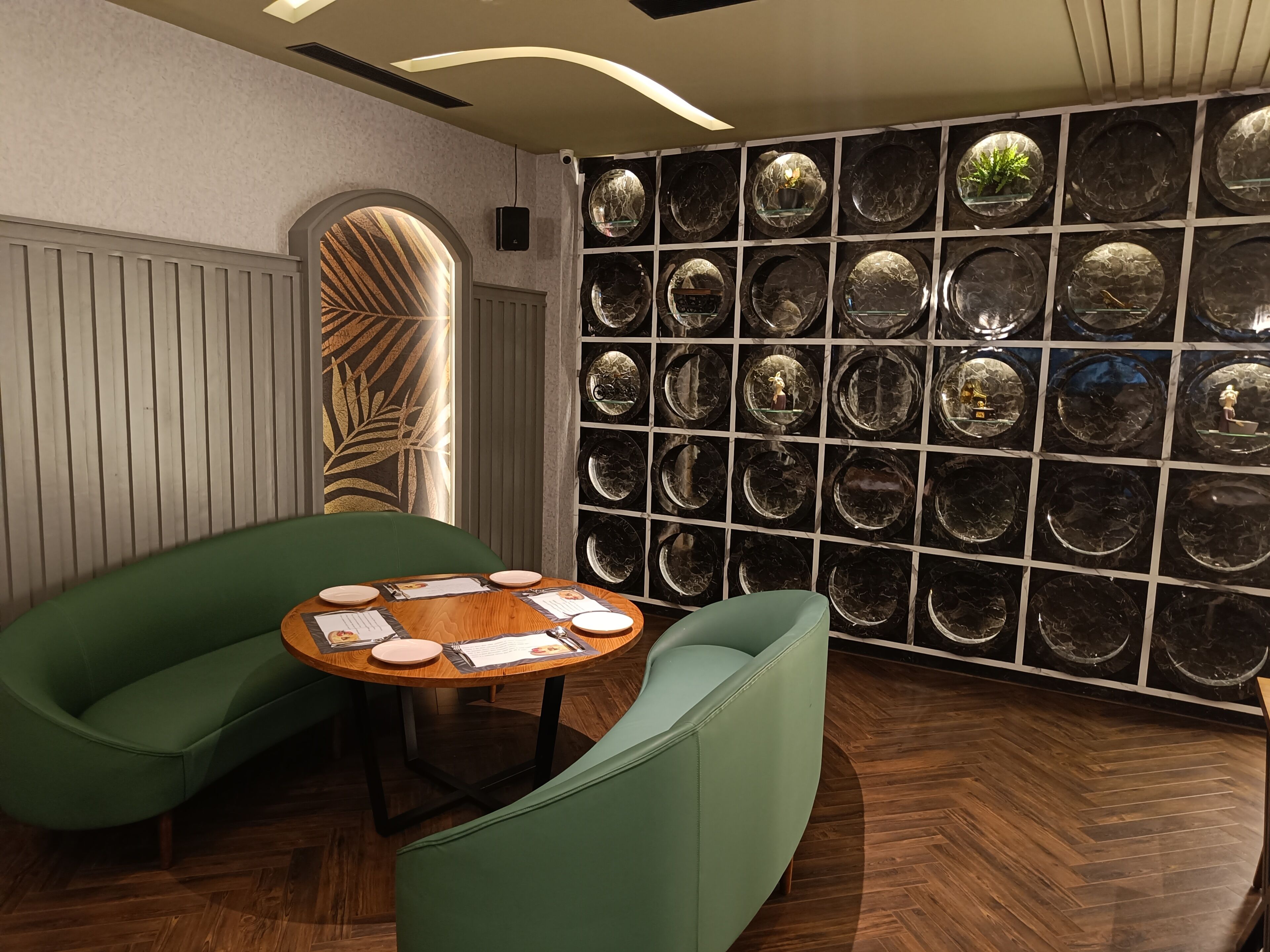 A modern interior with a round wooden table set for dining, two green upholstered chairs, and a wall of circular black marble tiles with various items displayed.