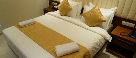 A neatly made bed with a white and gold bedspread, two gold pillows, and a white towel folded on the bed. There is a wooden nightstand with a telephone and a green box beside the bed.
