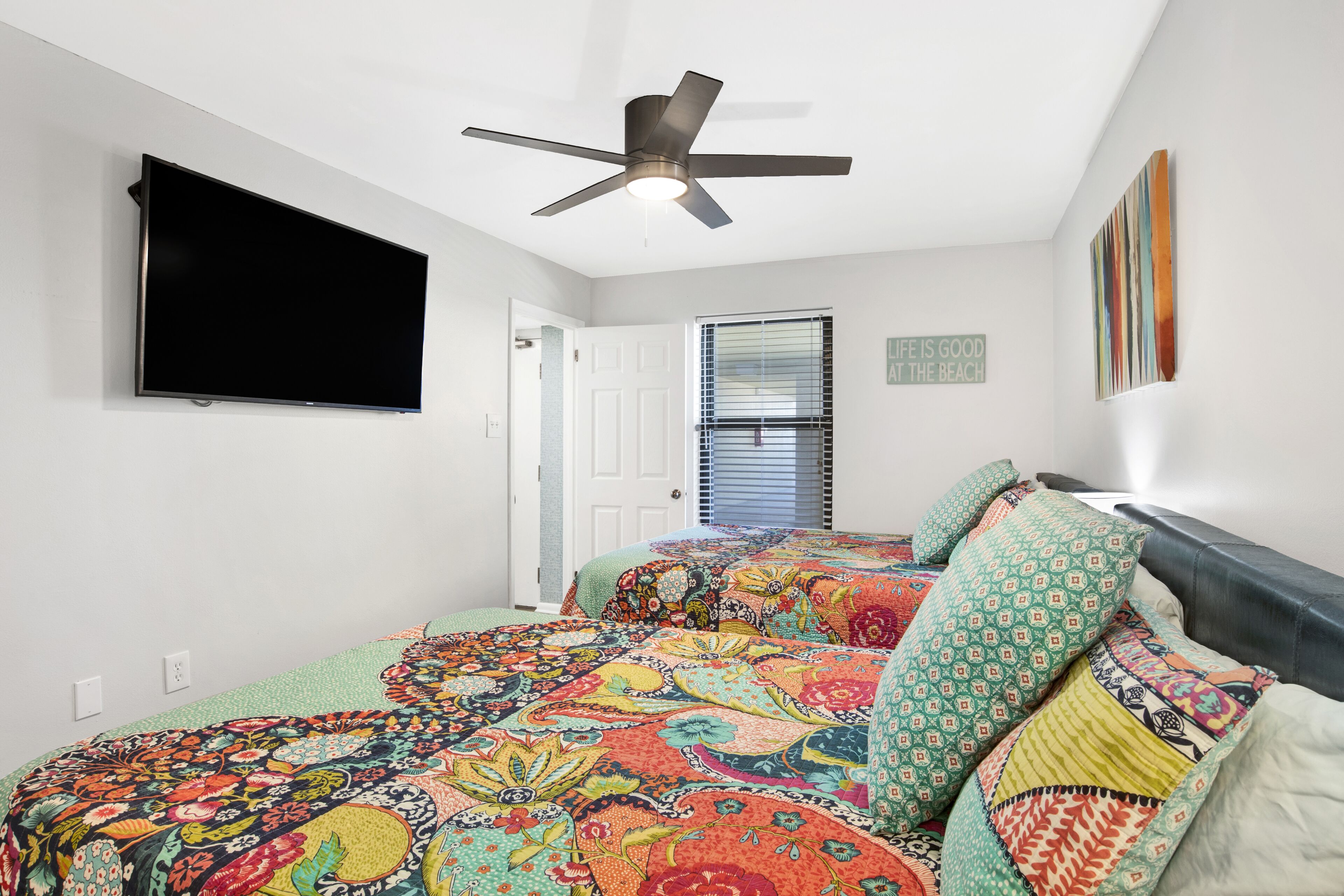 A bedroom with a bed, a ceiling fan, a TV mounted on the wall, a window with blinds, and a door.
