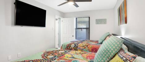 A bedroom with a bed, a ceiling fan, a TV mounted on the wall, a window with blinds, and a door.