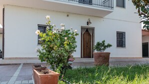 Un edificio con fachada blanca, una puerta de madera y un balcón. Hay plantas en macetas y un banco de madera en primer plano.