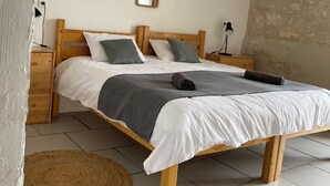 Un lit bien fait, avec une couverture et des oreillers gris, une tête de lit en bois, une table de chevet avec une lampe, et un tapis rond en fibres naturelles sur un sol carrelé.