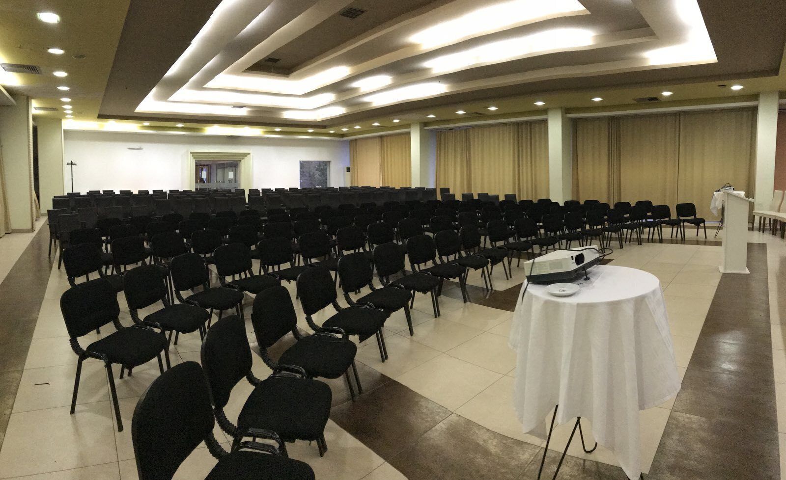 Une salle de conférence avec des rangées de chaises noires, une table blanche sur laquelle est posé un projecteur, et un pupitre.
