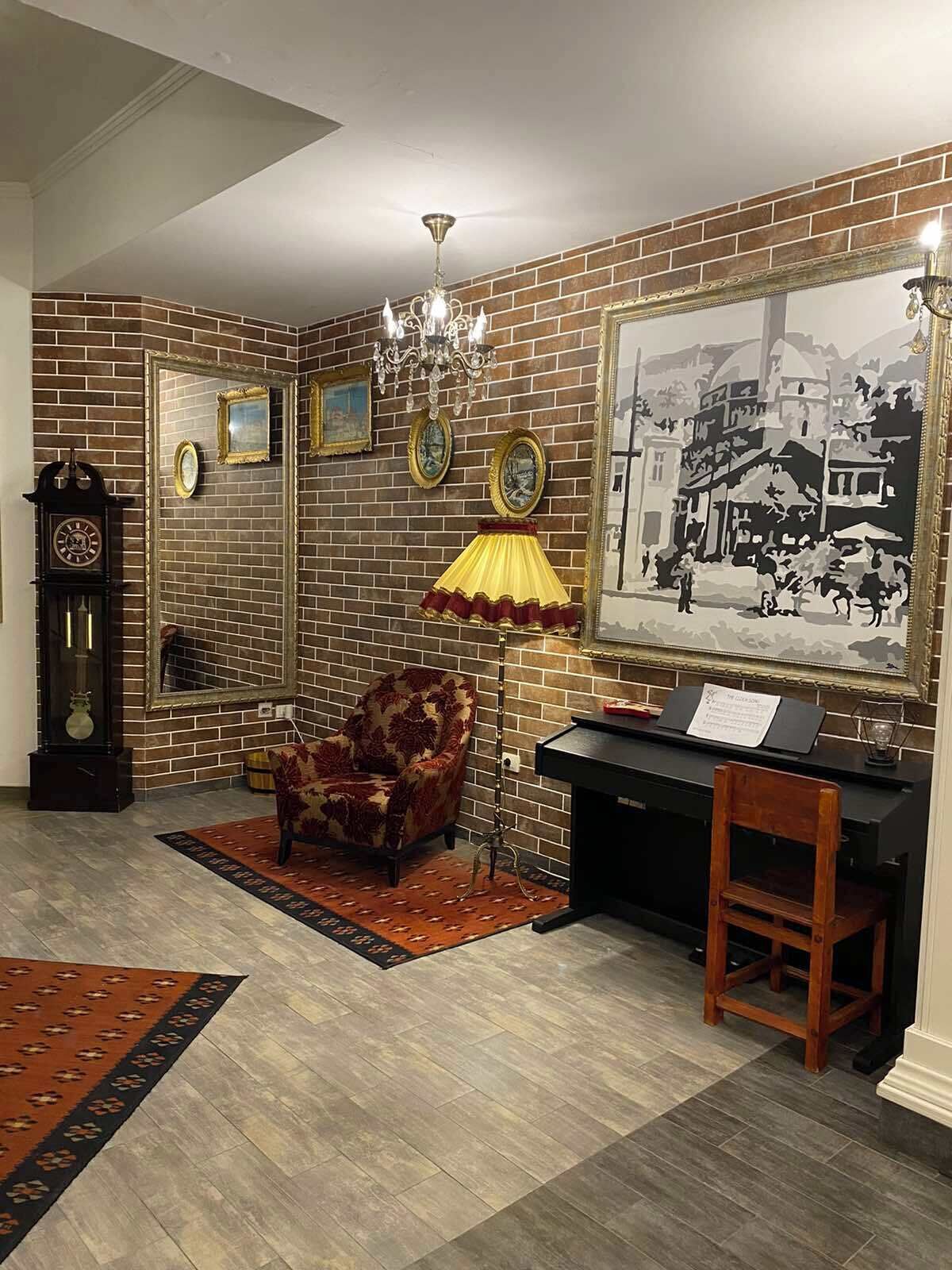 Una habitación con pared de ladrillo, un candelabro, un cuadro grande enmarcado, un reloj, una silla y un escritorio con lámpara.