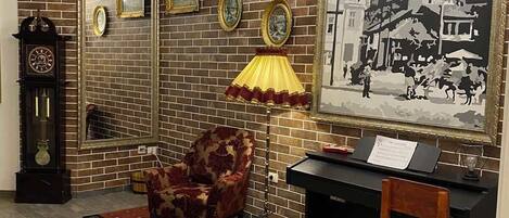 Una habitación con pared de ladrillo, un candelabro, un cuadro grande enmarcado, un reloj, una silla y un escritorio con lámpara.