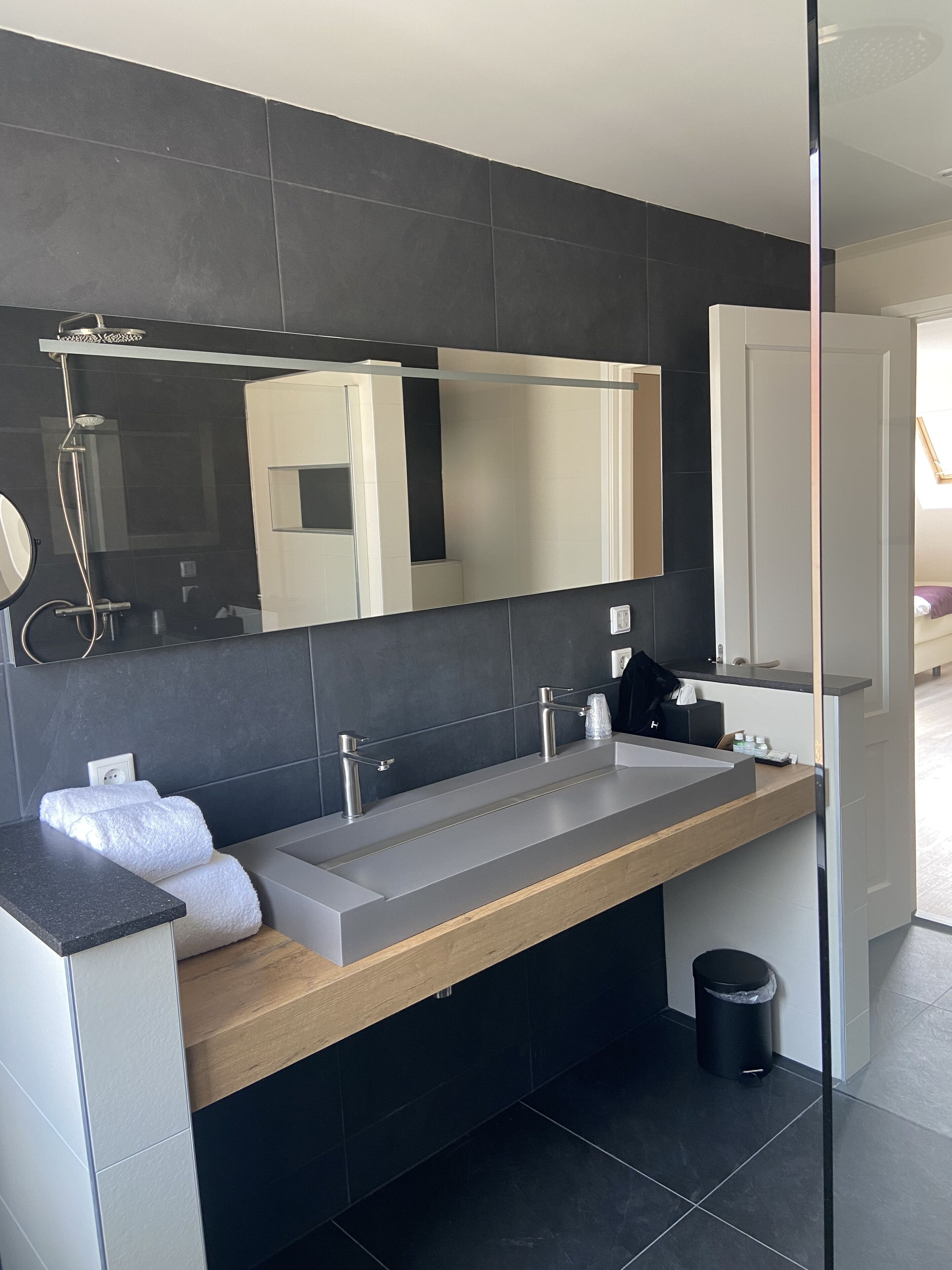 A modern bathroom with a large mirror, a wooden vanity, a shower, and a black trash bin.
