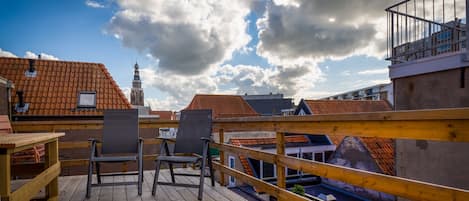 Een houten terras met stoelen en een tafel, met uitzicht op een stadsbeeld met rode daken en een toren.