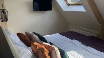 Un dormitorio con techo inclinado, televisor montado en la pared, cama con muchas almohadas y tragaluz.