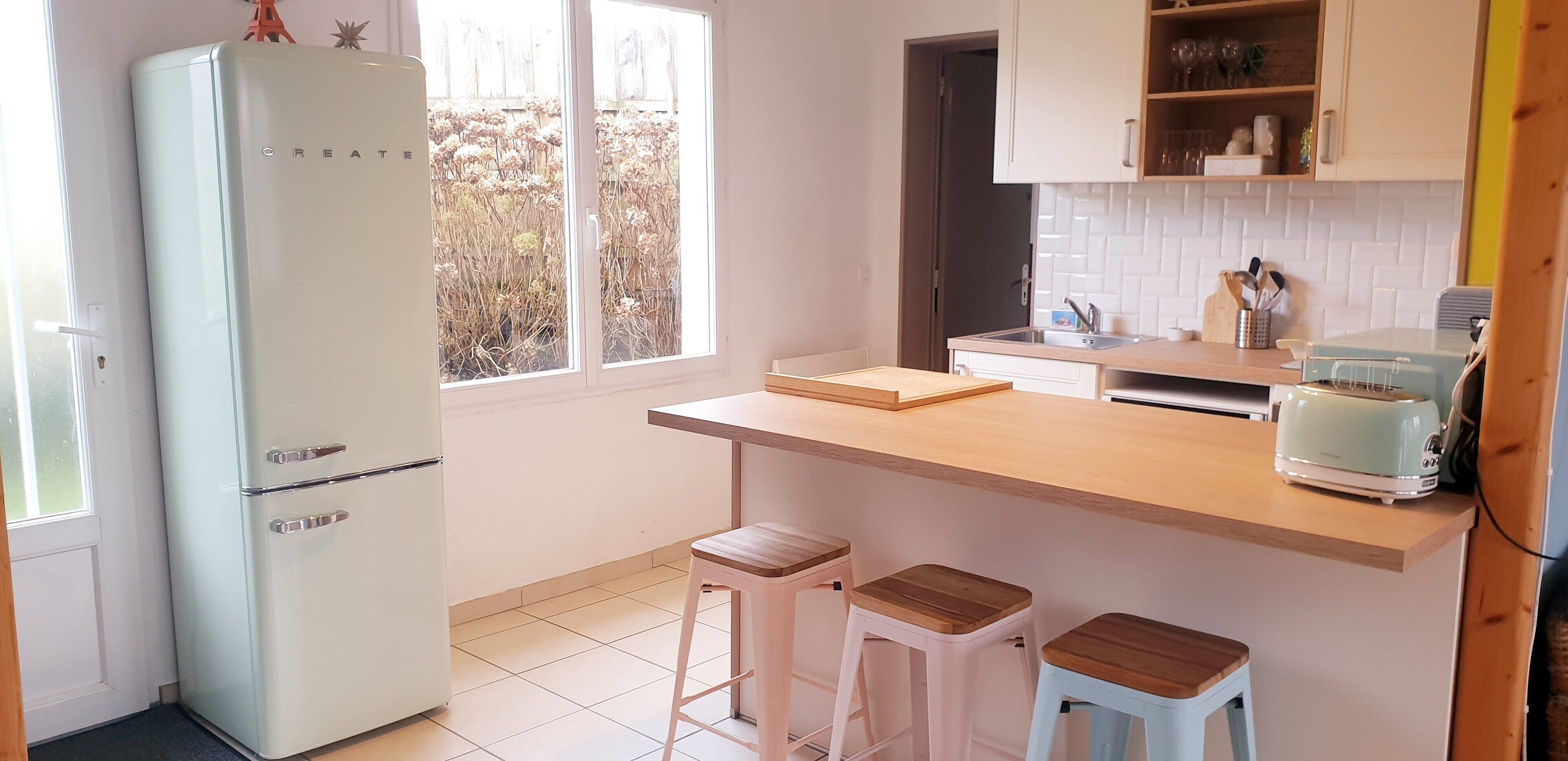 A compact kitchen with a white refrigerator, wooden countertop, two stools, and a toaster.